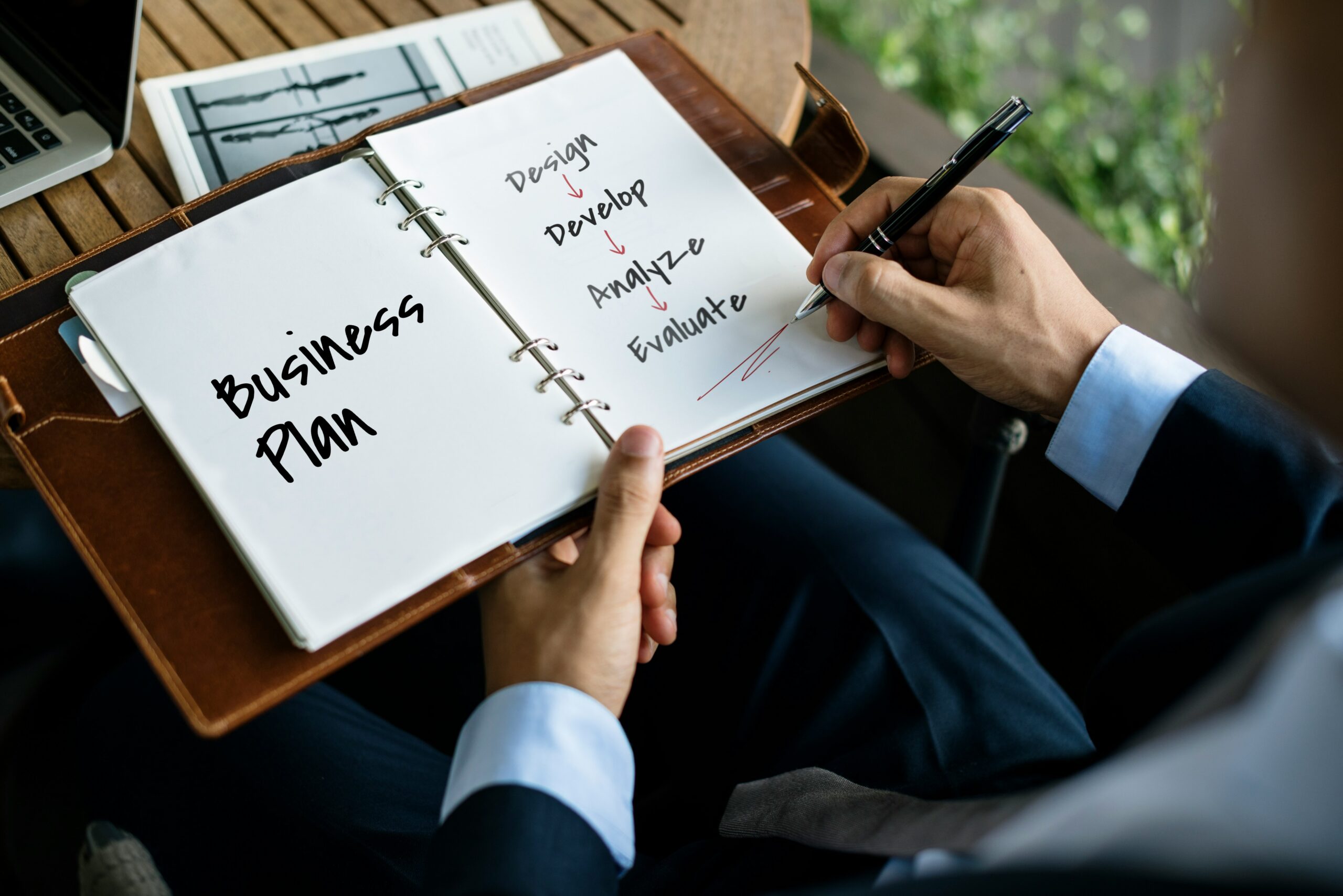 Hand writing in a planner showing a business plan flow: Design → Develop → Analyze → Evaluate with red arrows on the page.