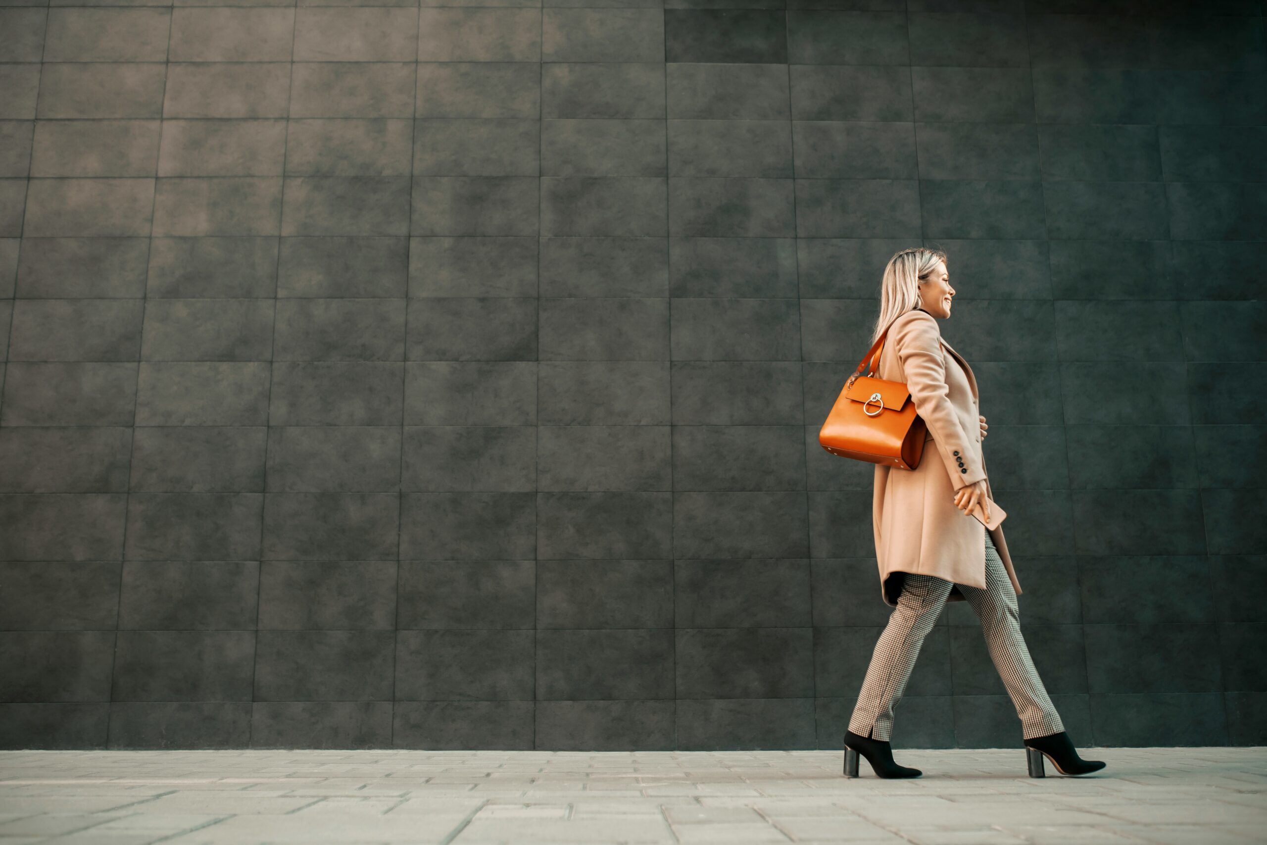 Side view of a woman in a beige coat carrying an orange handbag, walking past a dark gray tiled wall.