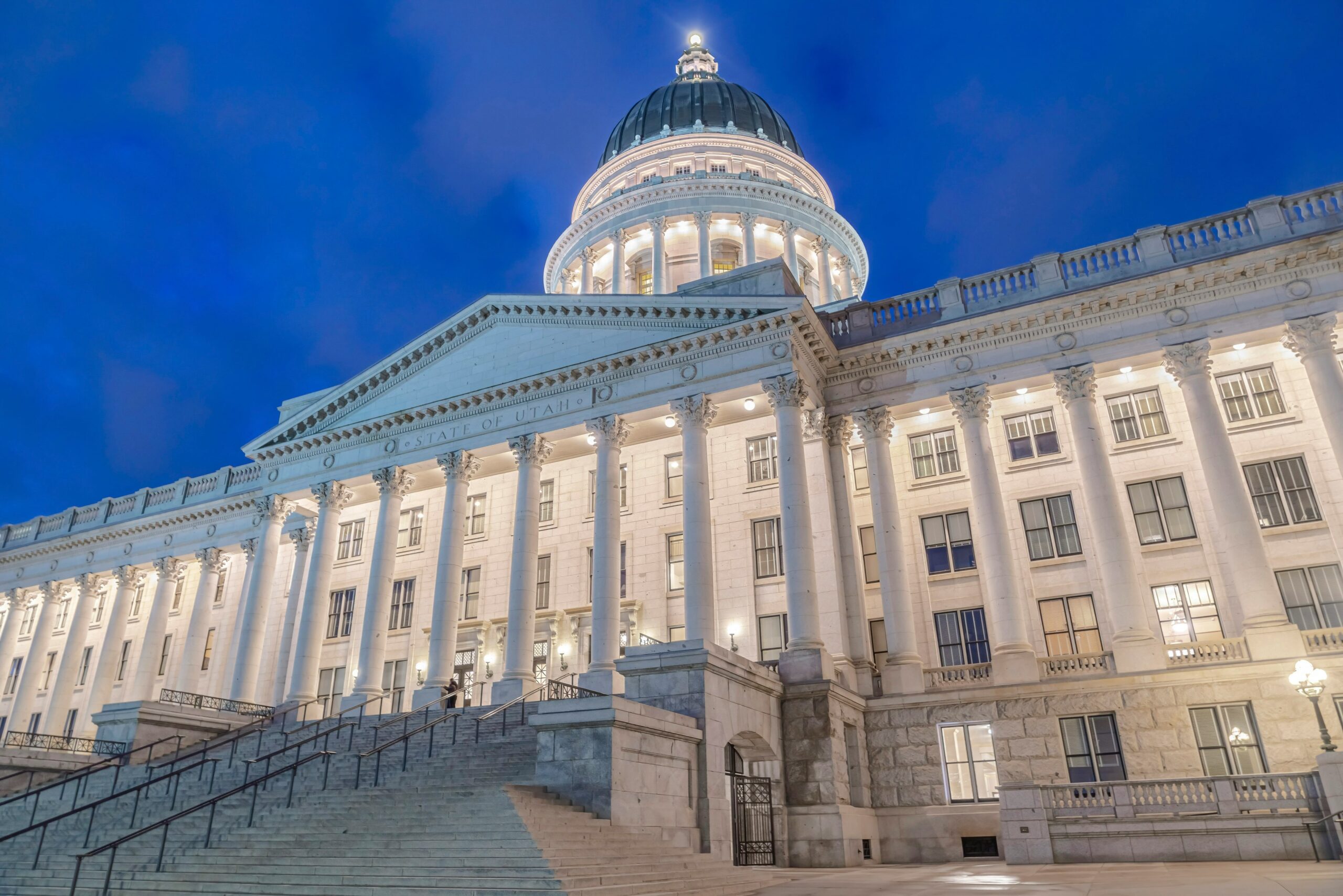 Front view of a neoclassical government building with a large illuminated dome, tall columns, and broad stone stairs at dusk.