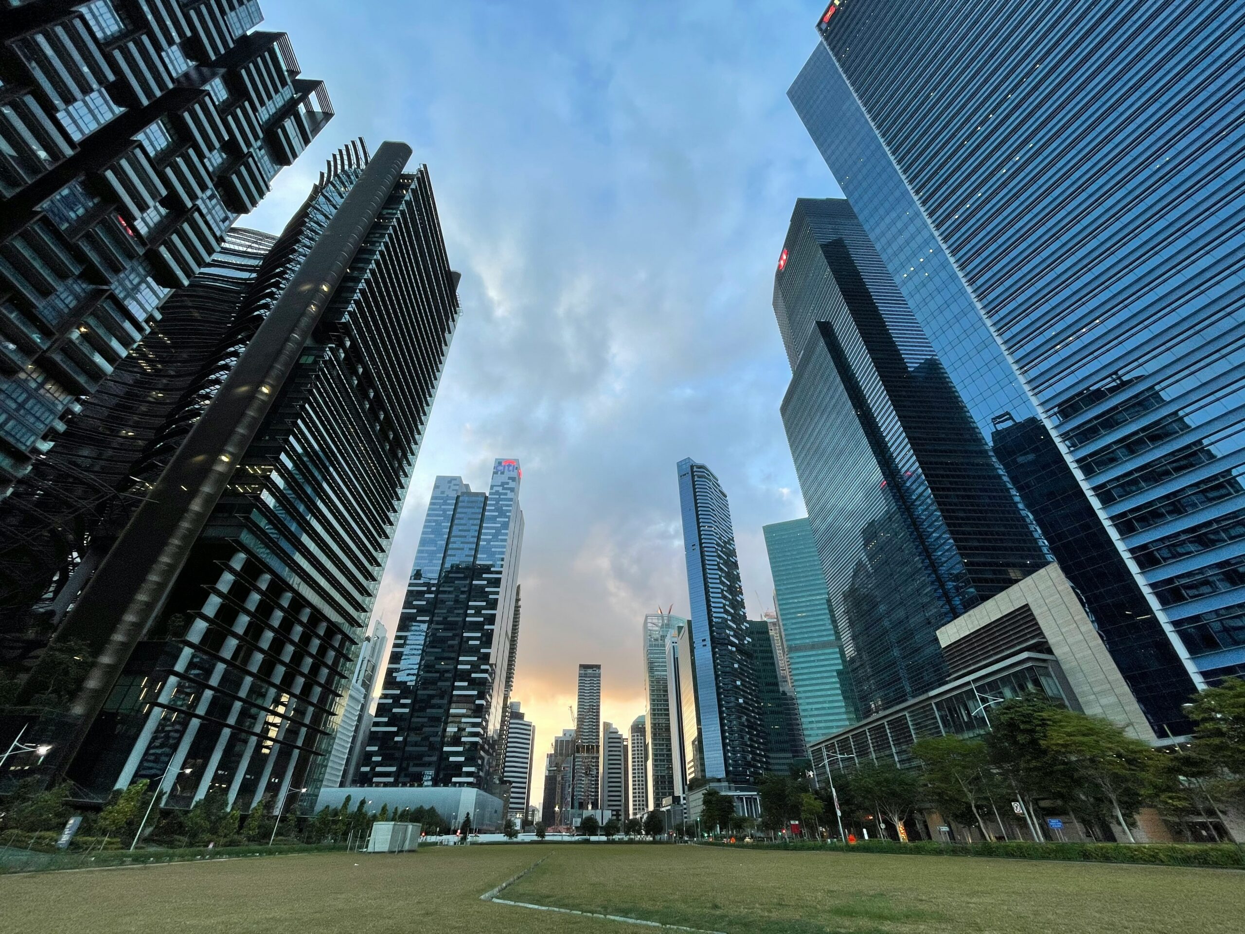 Skyscrapers frame a grassy plaza under a blue cloudy sky, seen from ground level looking up between tall glass buildings.