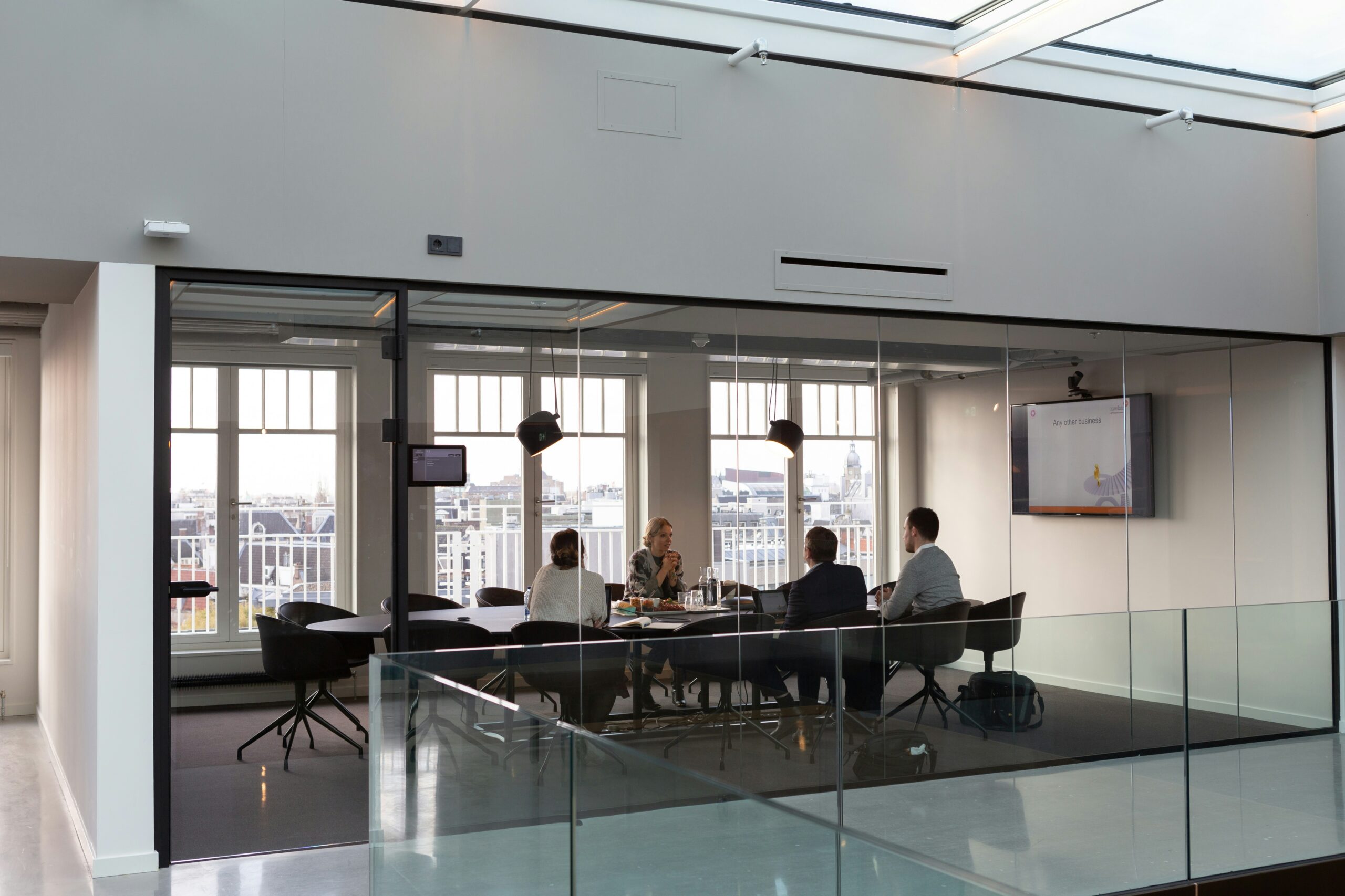 rodeo-project-management-software-ONe-snuCaqQ-unsplash - AEGIS LAW Group of businesspeople seated around a glass-walled conference table in a bright, modern office with a skylight, city skyline beyond.