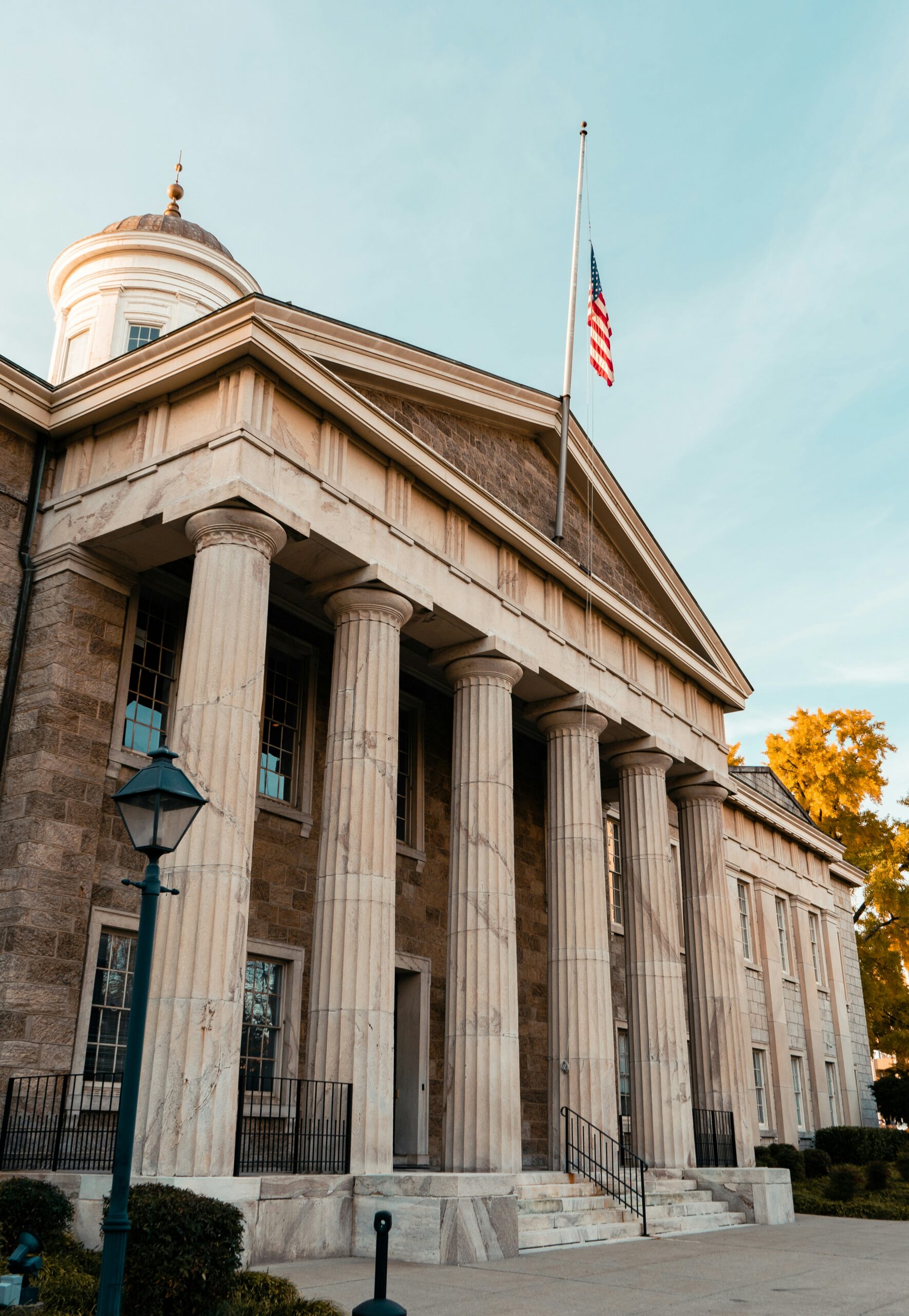 Front view of a neoclassical stone building with tall columns, steps, and an American flag flying from a flagpole against a clear blue sky.