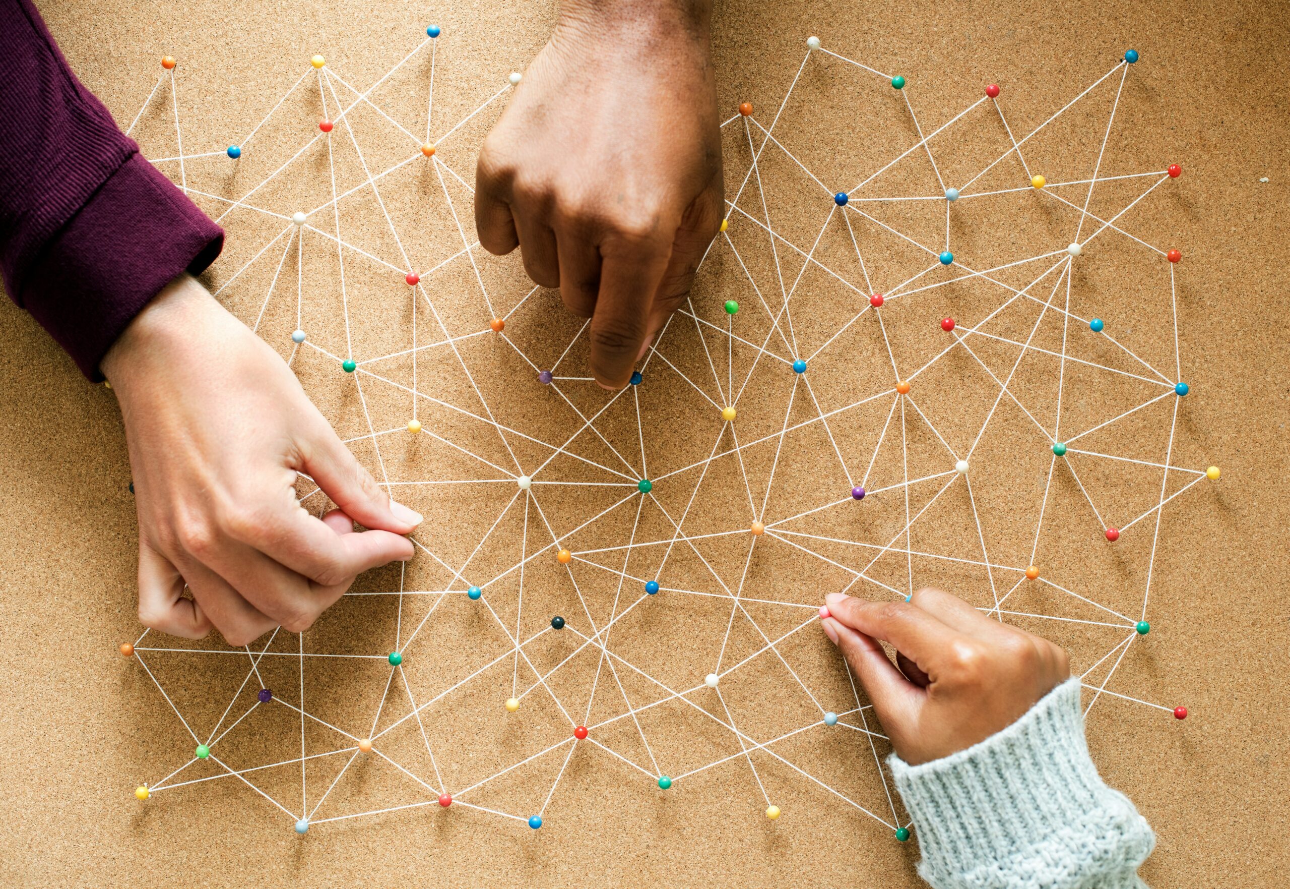 Hands place colored pins and string on a corkboard to form a network diagram, symbolizing collaboration and connections.