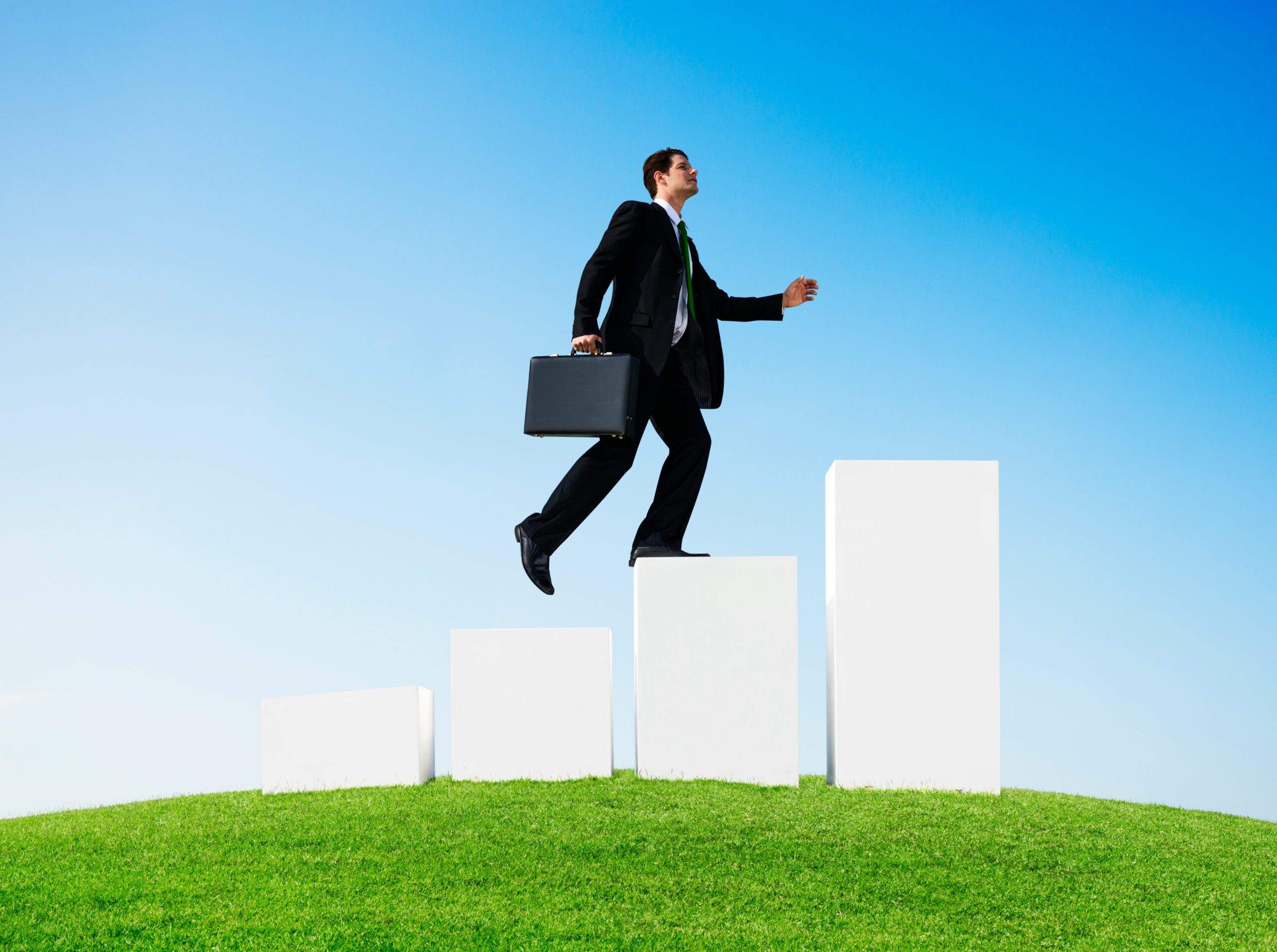 Businessman in a dark suit with a briefcase steps up white rectangular blocks against a bright blue sky and green grass.