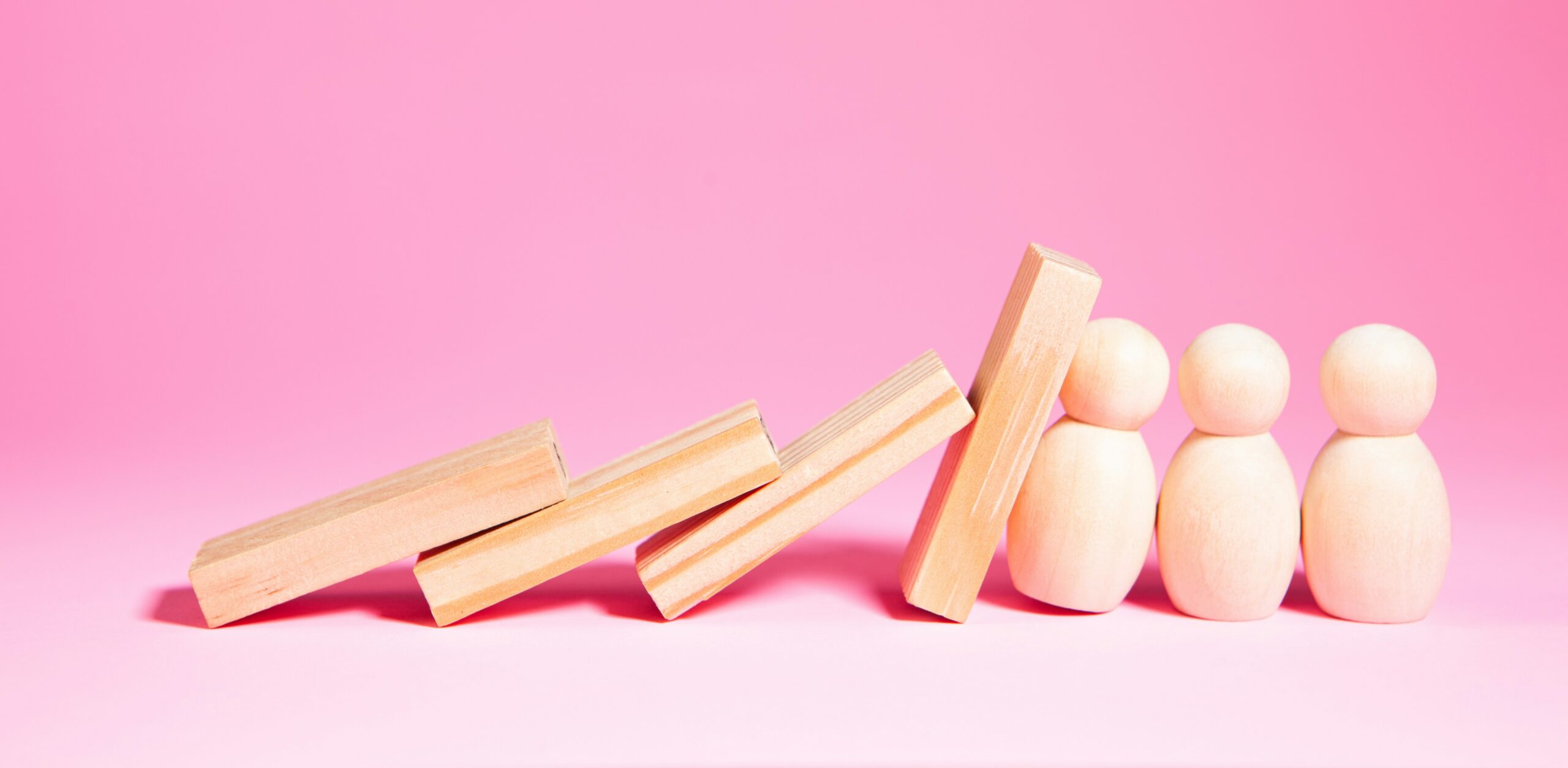 Wooden peg dolls stand as wooden blocks lean and topple toward them on a pink background.