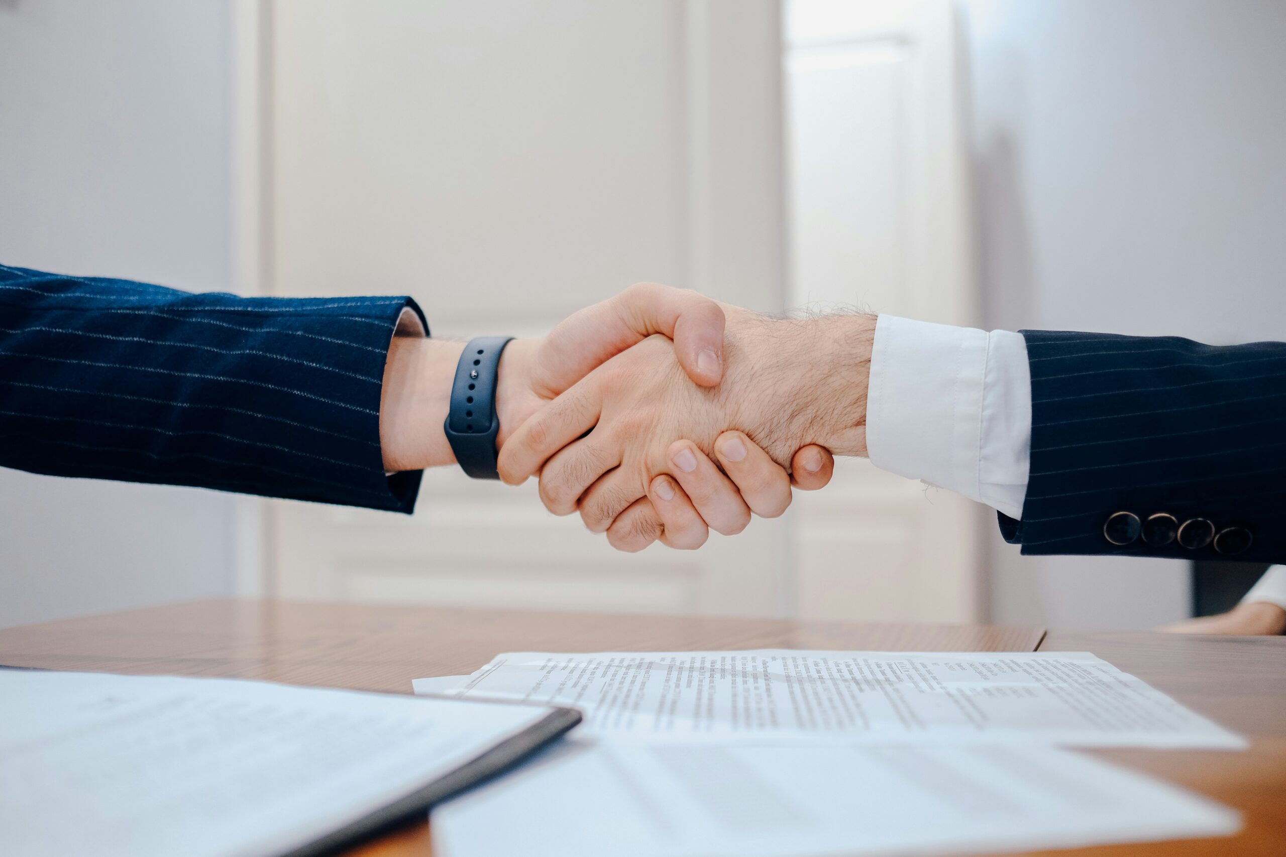 Two professionals in suits shake hands over a desk covered with documents, signaling an agreement.