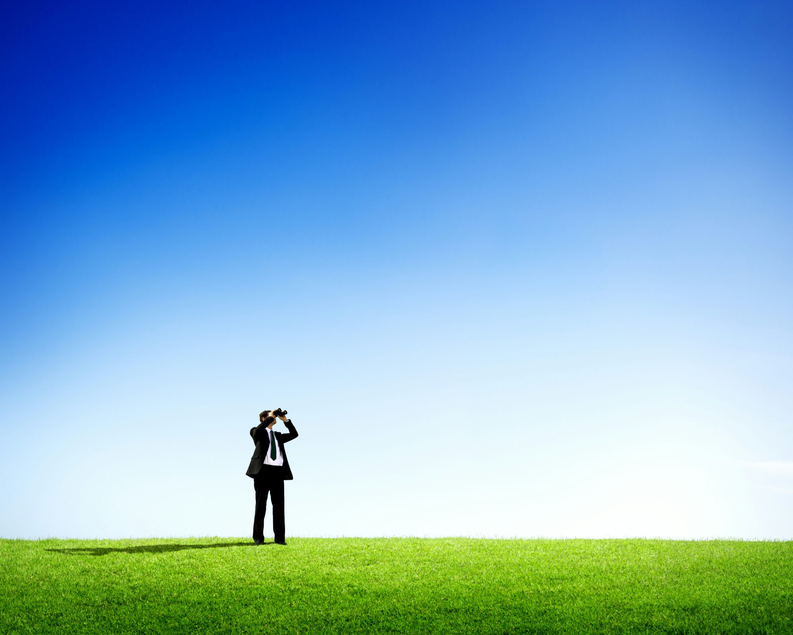 Two people in suits standing apart on a grassy hill, looking through binoculars toward the sky.