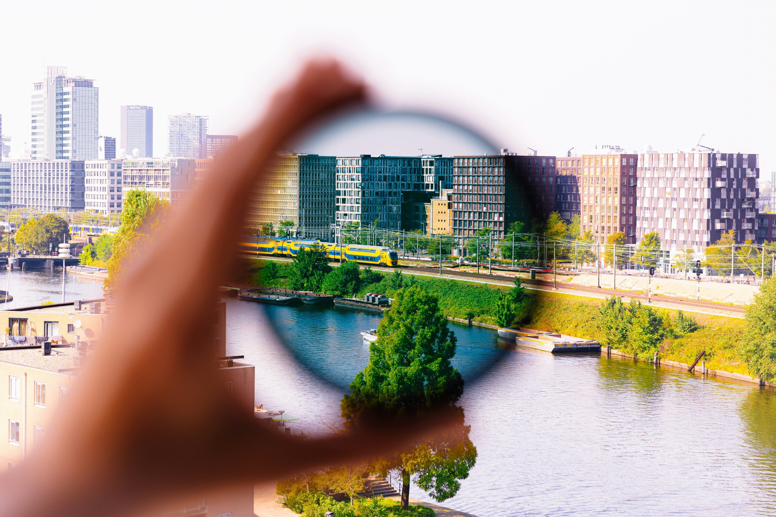 A hand holding a circular lens that frames a city skyline with a river and a yellow train passing by.