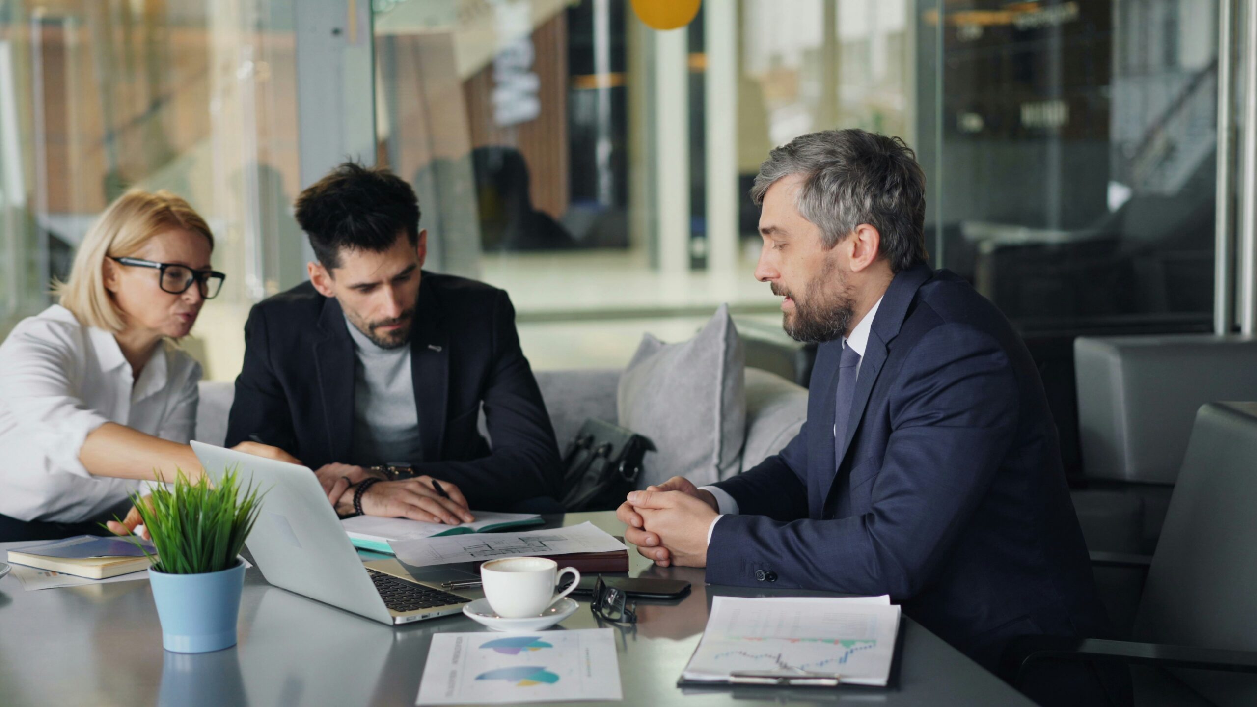 Three professionals in a business meeting around a table with laptops, papers, and charts, actively discussing.