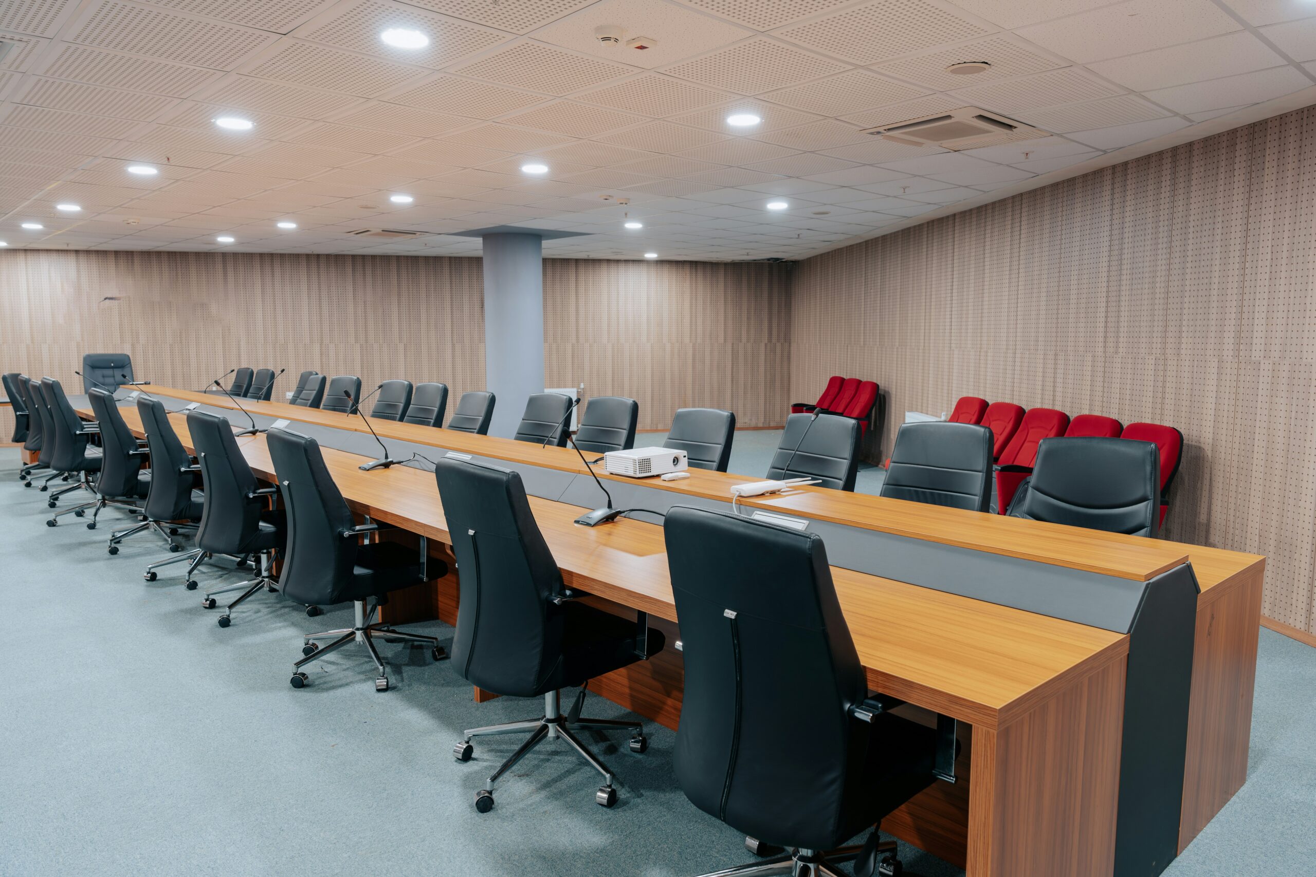 Modern boardroom with a long wooden table and black leather chairs, red seating along the back wall, and bright ceiling lights.
