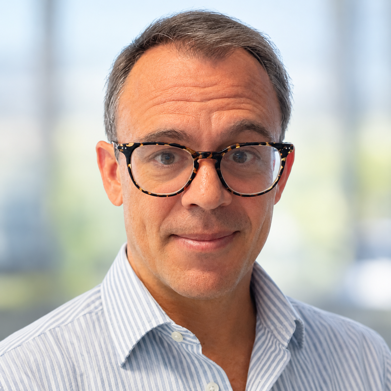 Portrait of a middle-aged man with tortoiseshell glasses and a blue striped shirt, looking at the camera in a bright office.