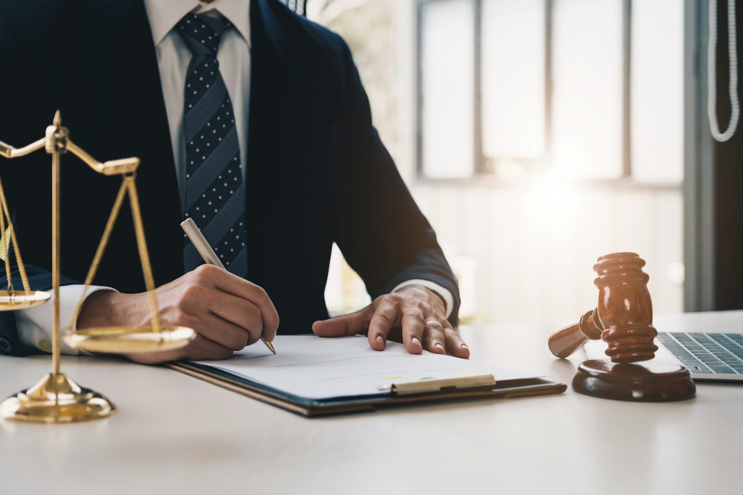 Lawyer in a suit signing papers at a desk with scales of justice and a gavel nearby.