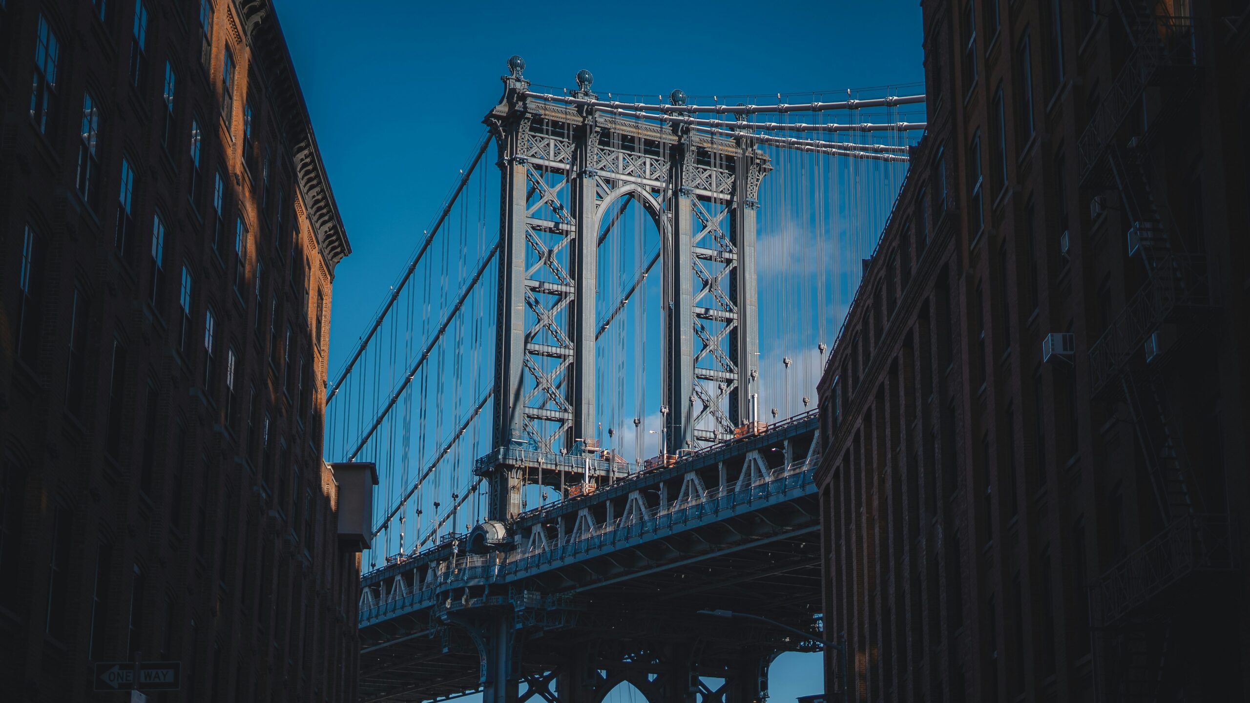 Steel suspension bridge framed by brick buildings on either side, cables spanning the skyward towers against a bright blue sky.