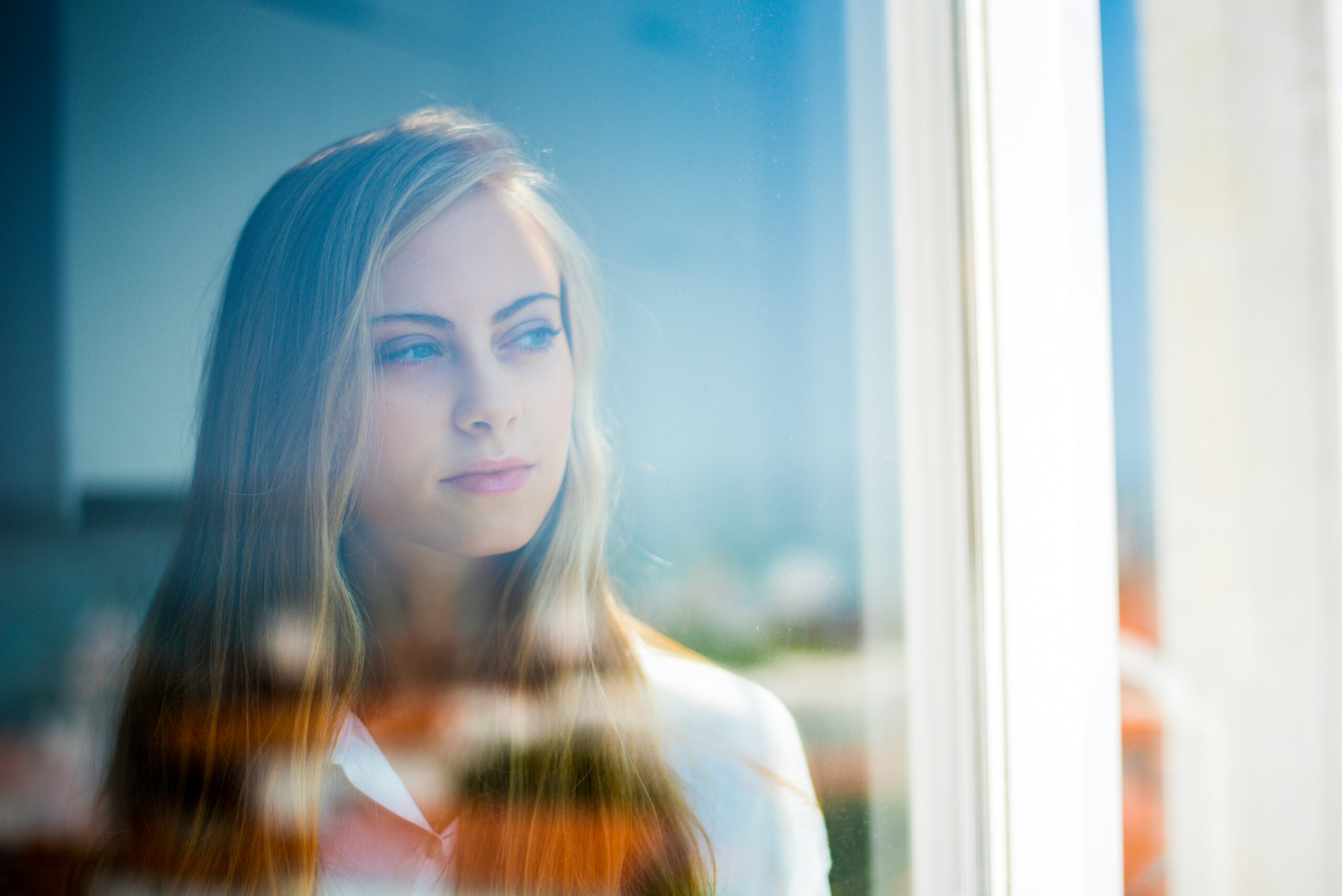 Woman gazing out a glass window, with reflections creating a soft, moody blur around her face.